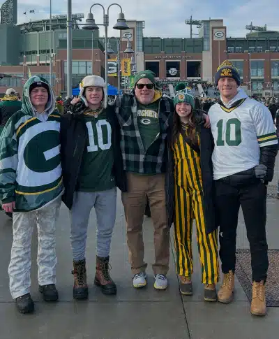 Mobile_Meet_the_Doctor_One_of_1 Five people wearing Green Bay Packers gear and winter clothing smile and pose together outside Lambeau Field, with the stadium and other fans visible in the background.